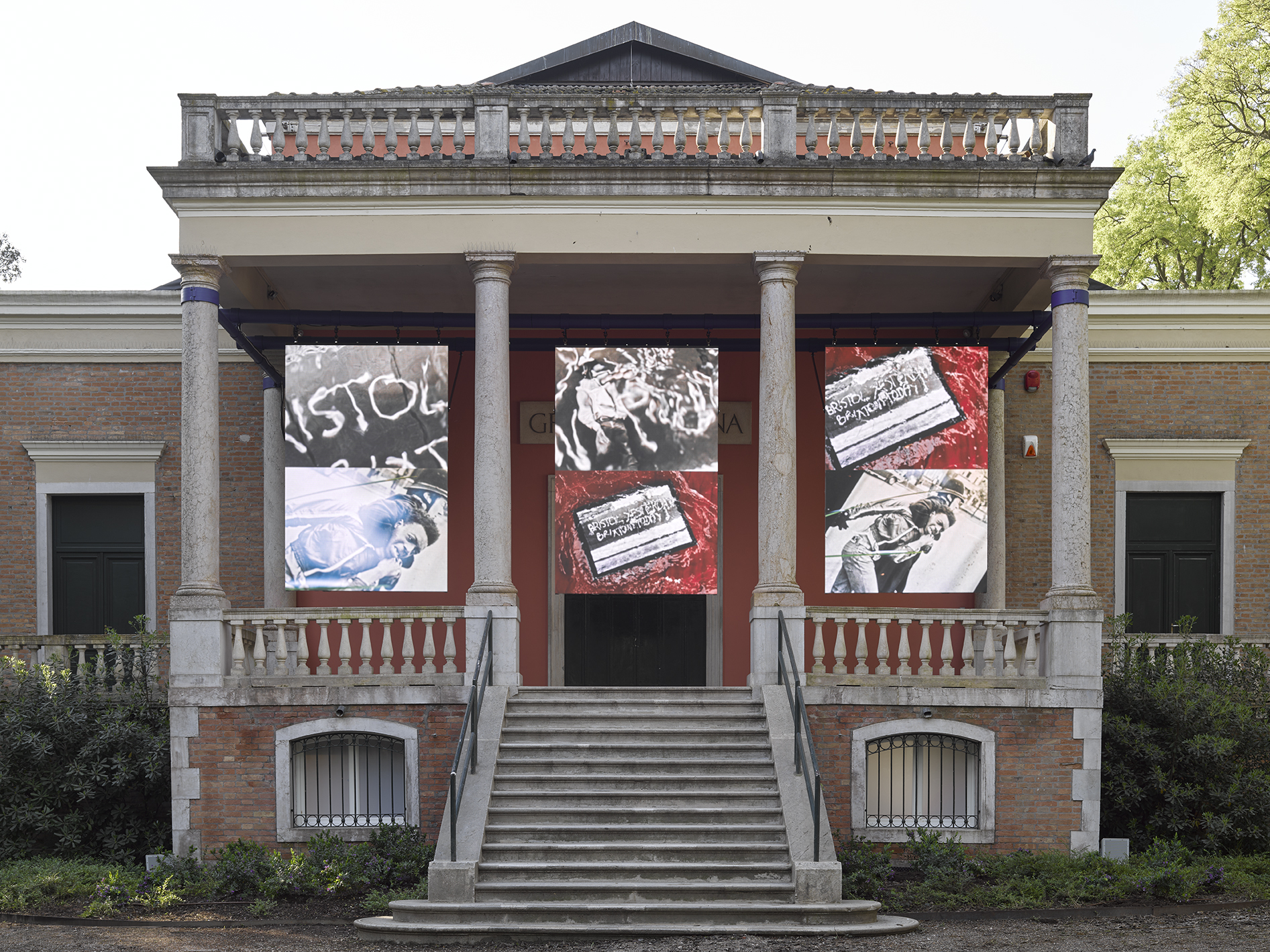 John Akomfrah- outside a large building with pillars and a staircase to the main door and red, white and black graffiti style painting hung on the exterior 