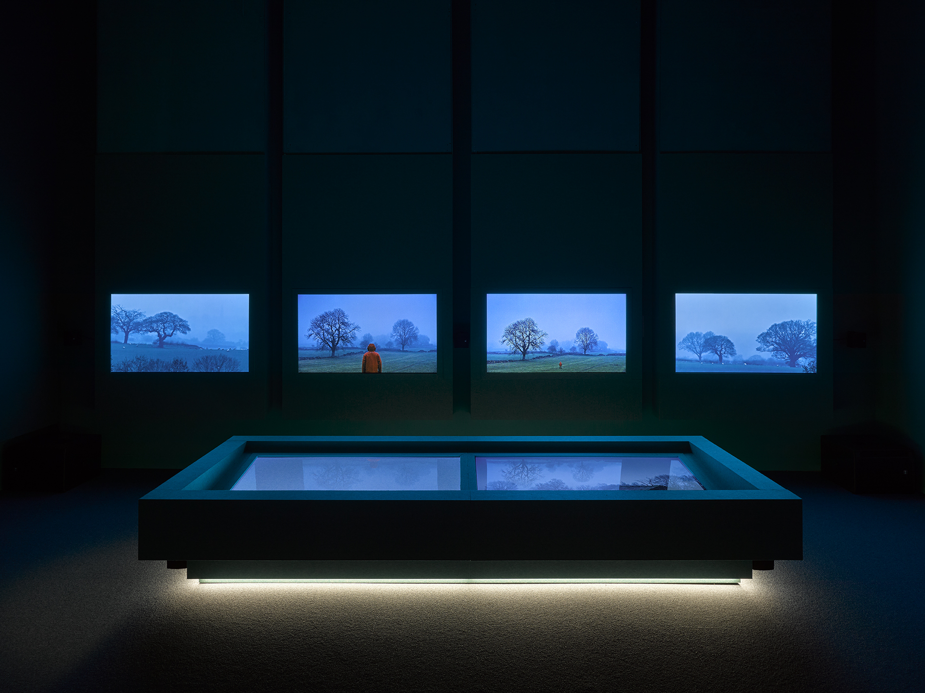 John Akomfrah - blue screens in a room with a blue lit up table in the middle of a dark room