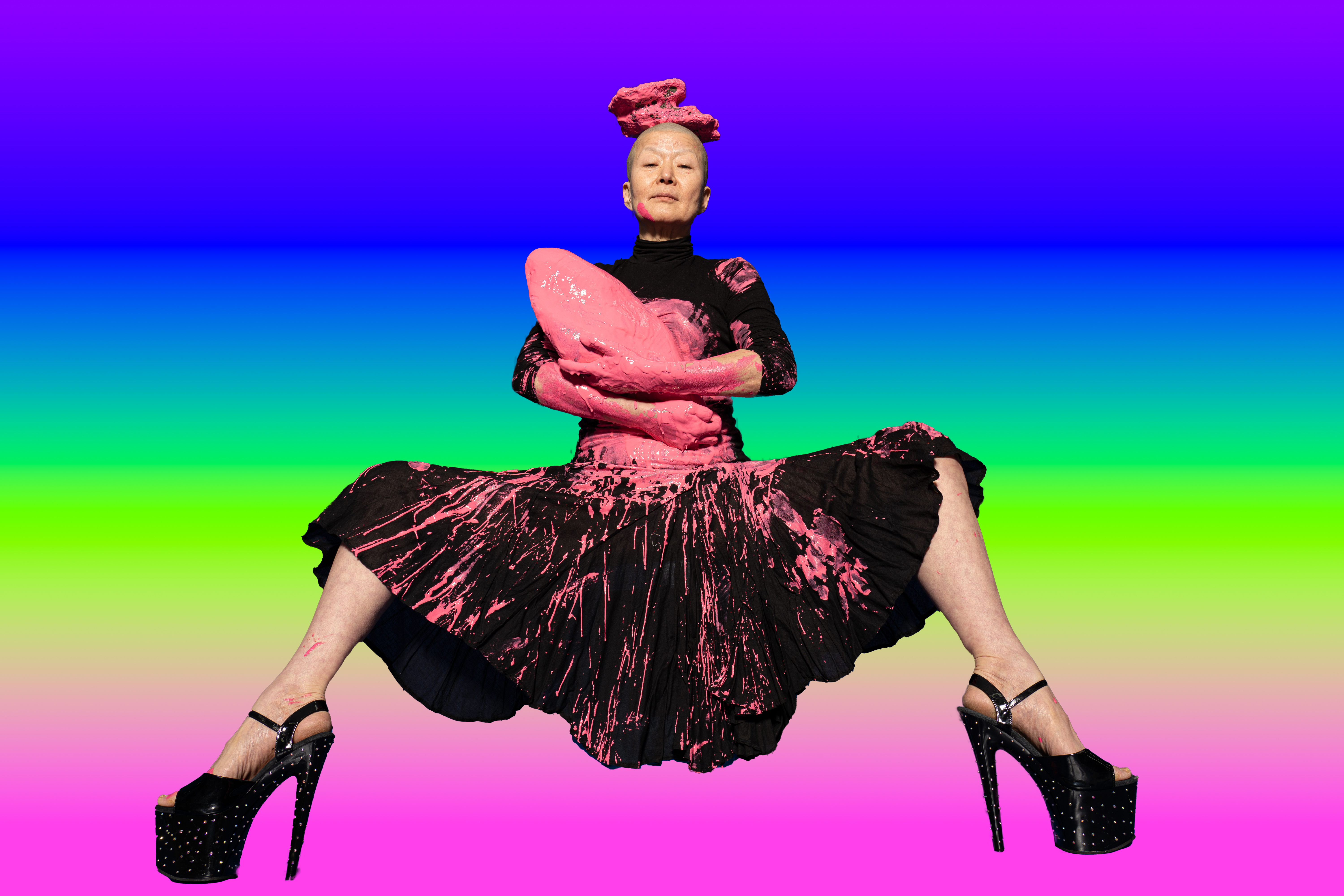 Eun-Me Ahn - sitting on a rainbow background wearing platform heels and a black dress with pink paint all over her