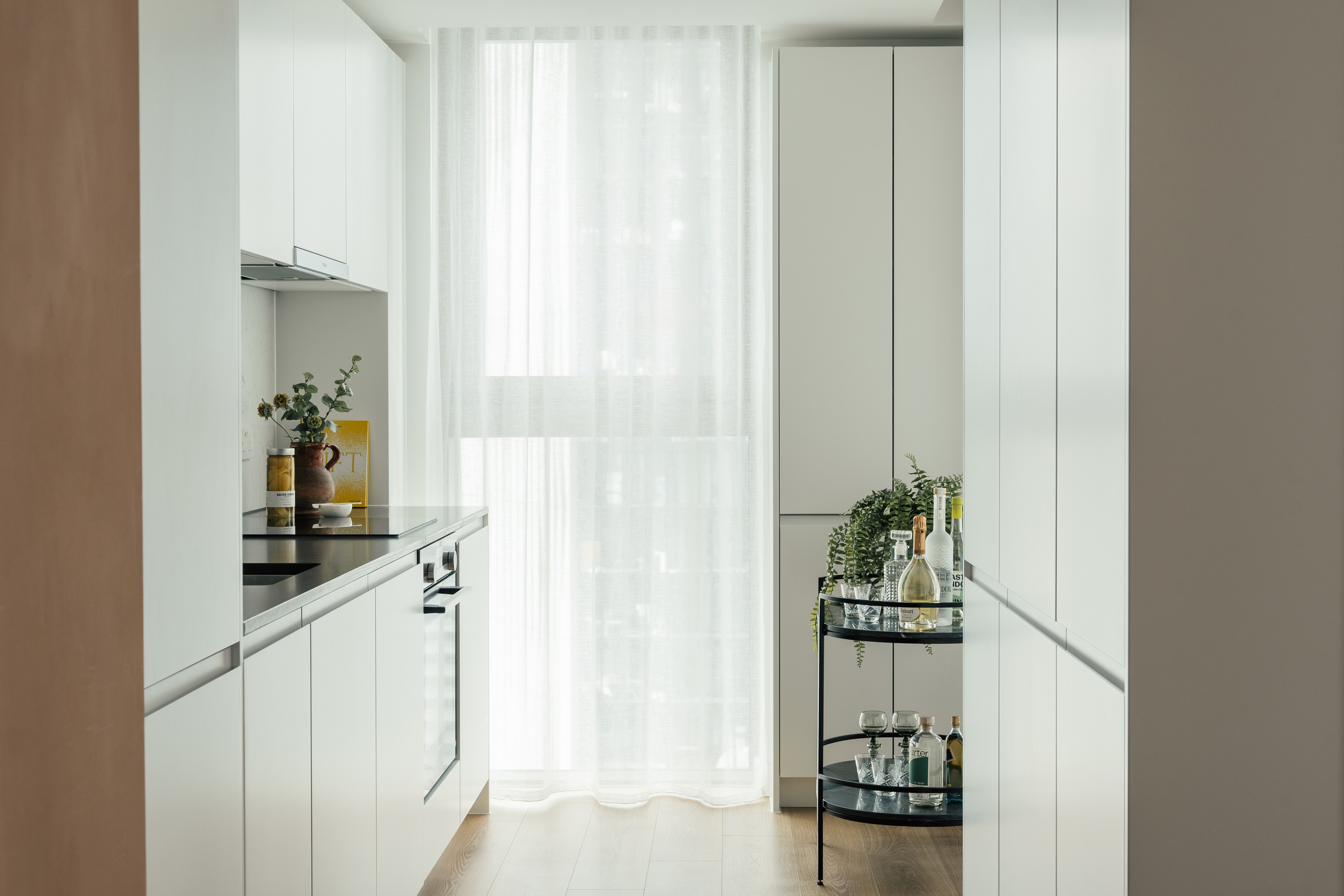 Property of the Week One Thames Quay - A modern kitchen at One Thames Quay with handleless white cabinetry, black countertops, and integrated appliances. A large window with sheer curtains and a bar cart with glassware and greenery are also featured.