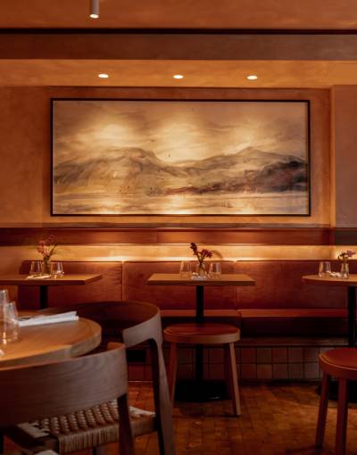 Art restaurants in London - warmly lit restaurant interior of Lolo in Bermondsey featuring wooden tables and cushioned bench seating. A large framed landscape painting serves as the focal point.